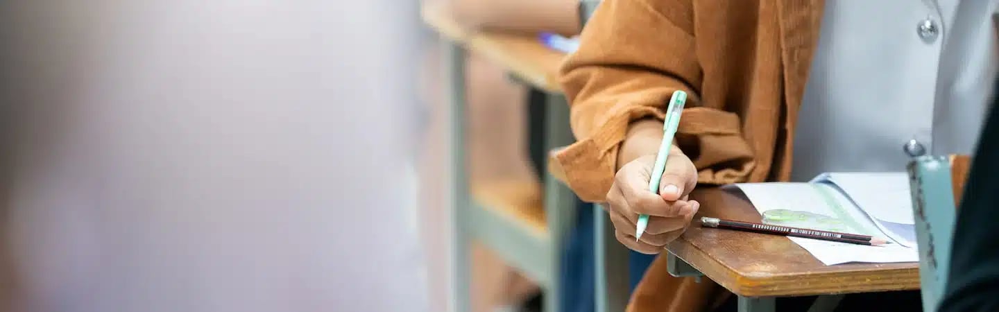 Student holding a pen over an open notebook on a desk in a classroom setting.