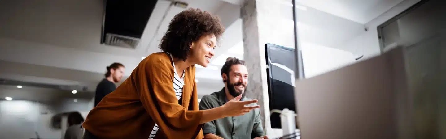 Two colleagues happily discussing work in a modern, open office space with computers and bright lighting.