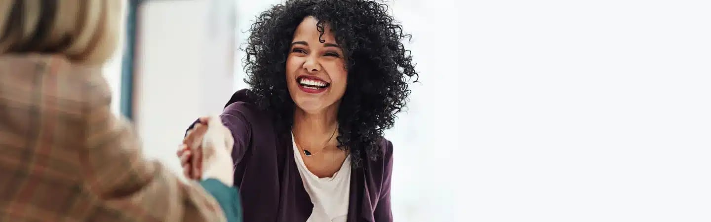 Woman with curly hair smiling and shaking hands with someone in a blurred background.