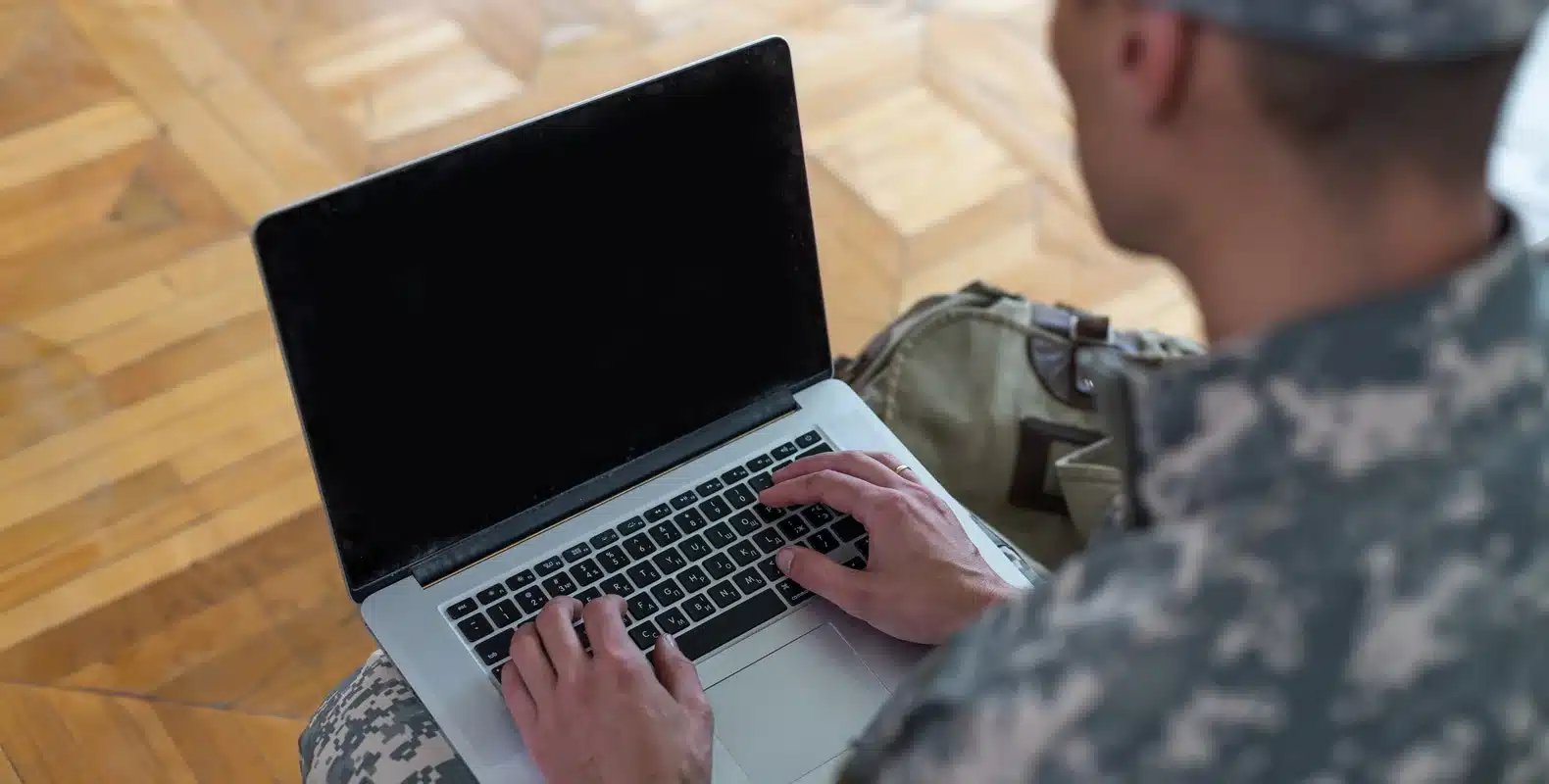 Person in military uniform typing on a laptop, sitting on a wooden floor with a backpack beside them.
