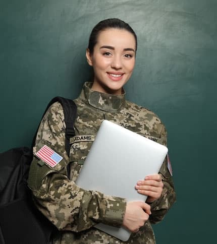 A woman in military uniform smiles while holding a laptop and a backpack. She stands in front of a dark green background. The uniform has an American flag patch and a name tag.