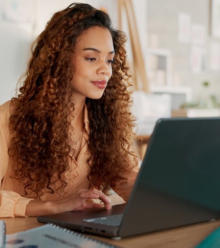 A woman with curly hair sits at a desk, typing on a laptop. She appears focused and is indoors, with notebooks and other items on the desk around her.