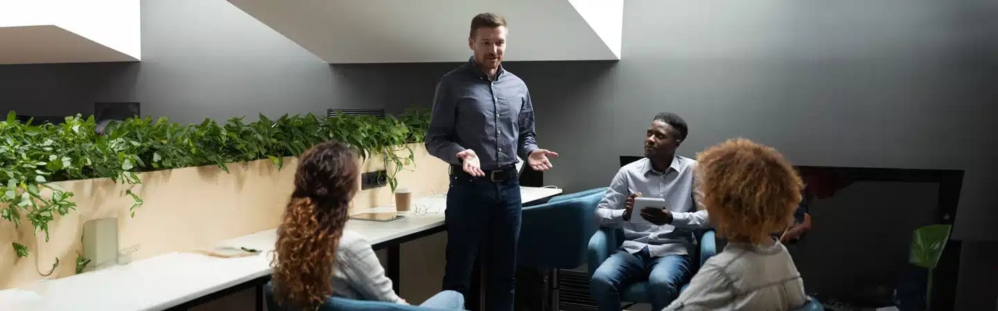 A man stands speaking to three seated people in a modern office with a plant-lined counter.
