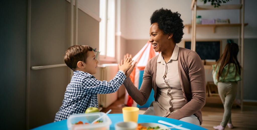 A young boy and an adult woman smile and give each other a high five while sitting at a table in a bright, cheerful room focused on early childhood education, with toys and a play tent in the background. Another child is playing nearby.