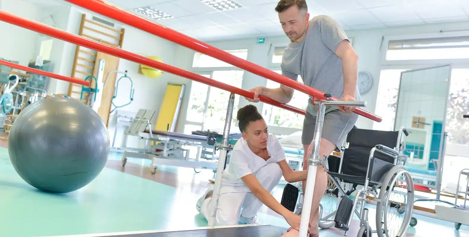 Occupational Therapy Assistant helps a man walk with parallel bars in a rehabilitation center, wheelchair and exercise equipment nearby.
