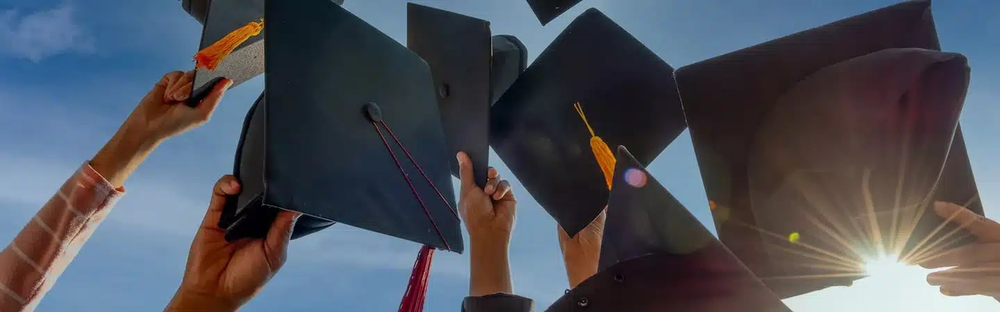 Hands raising graduation caps against a blue sky with the sun shining brightly in the background.