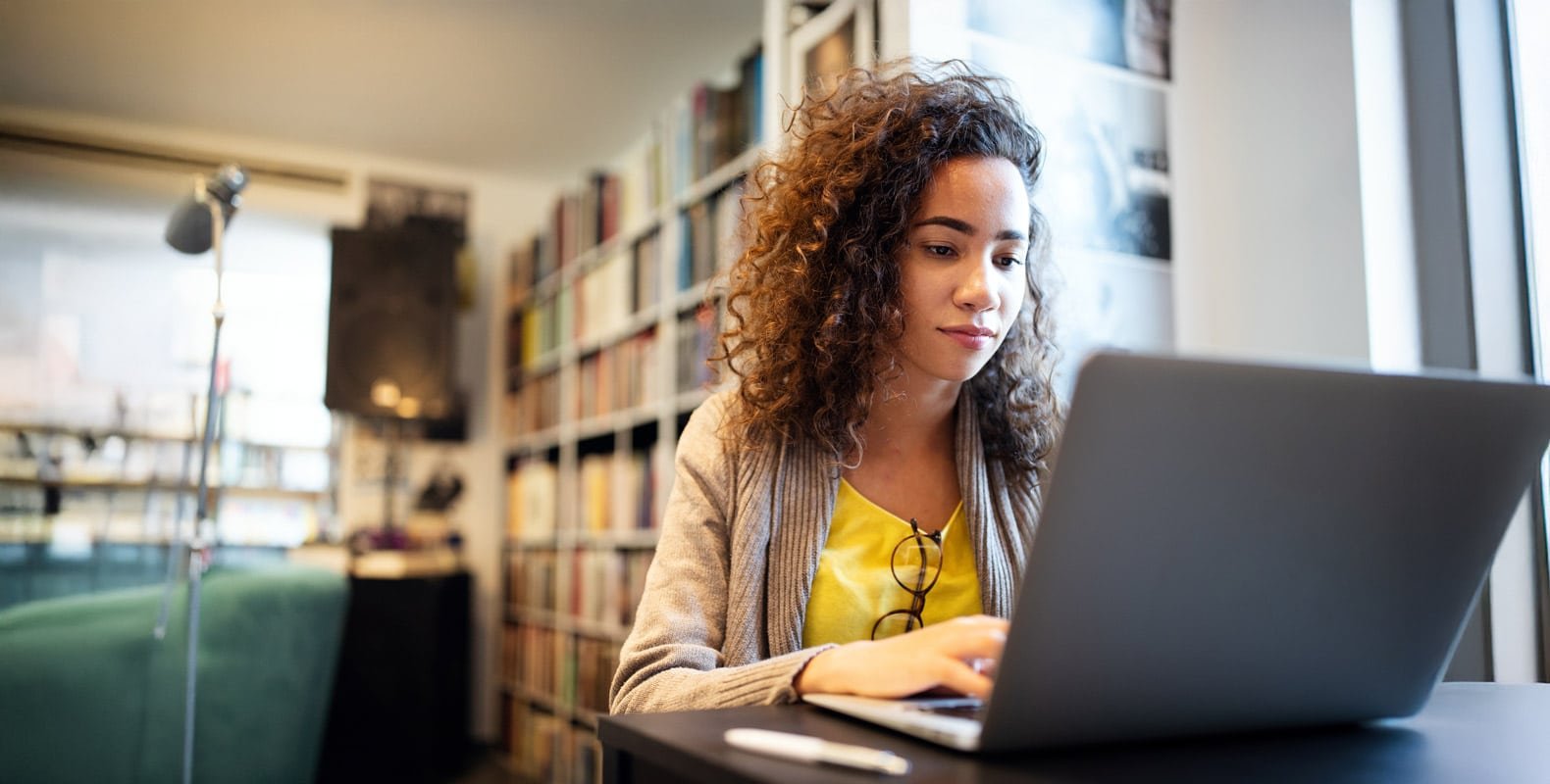 A person with curly hair in a yellow shirt and beige cardigan is sitting at a table using a laptop. They are focused on the screen. Bookshelves and a lamp are in the background, suggesting a library or study area.