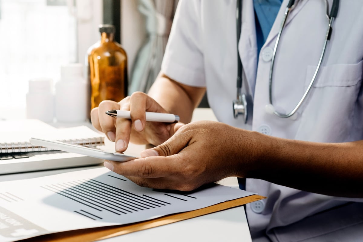 Doctor in white coat and stethoscope using a tablet and writing notes at a desk.