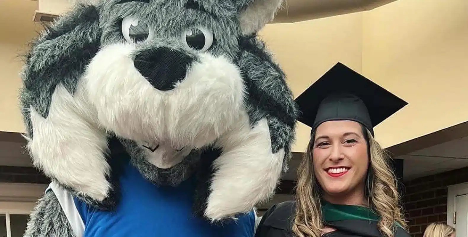 A person in a graduation cap and gown stands next to a large, friendly-looking wolf mascot indoors.