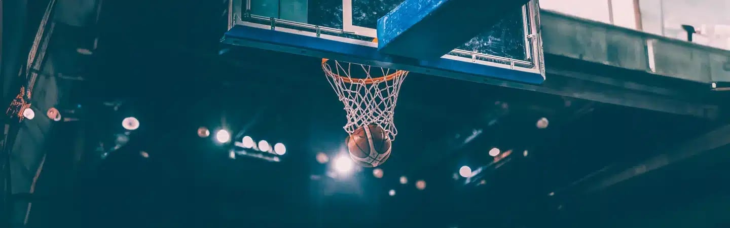 A basketball falling through the hoop on an indoor court, with stadium lights illuminating the background.