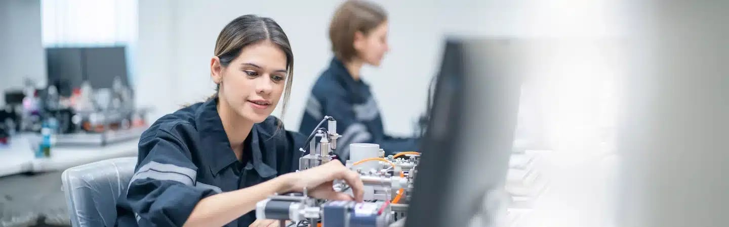 A woman working with electronic equipment in front of a computer in a lab environment, another person in the background.