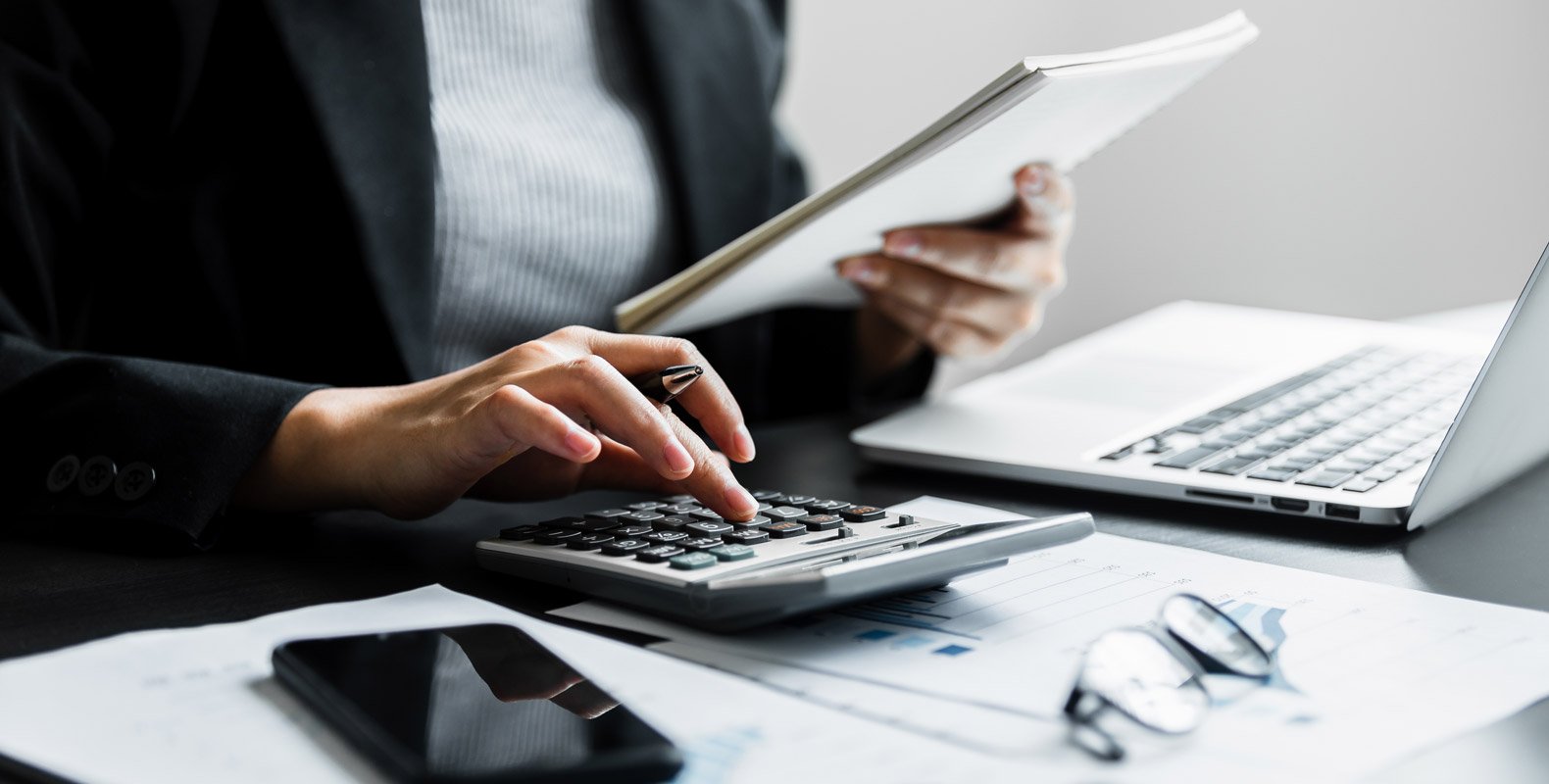 A person in business attire using a calculator, holding papers, with a laptop, documents, smartphone, and glasses on the desk, suggesting work related to a CPA or Accountant and highlighting the difference in their roles.