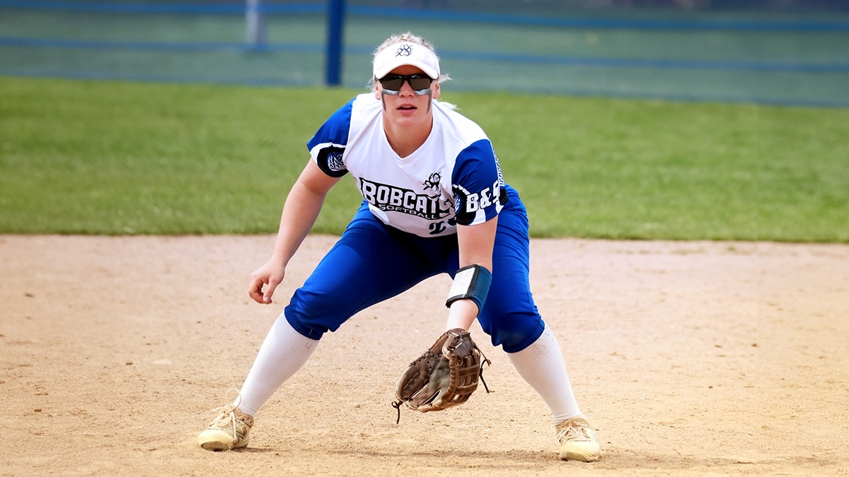 Softball player in a ready stance wearing a white and blue uniform and sunglasses, preparing to field a ball.