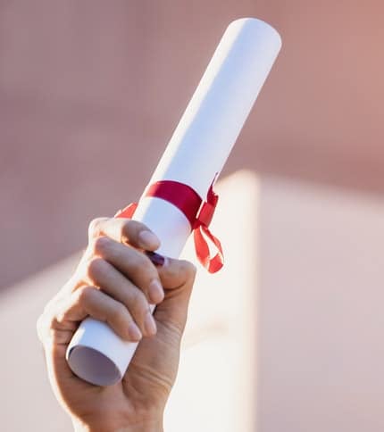 A hand holding a rolled-up diploma tied with a red ribbon, symbolizing achievement and academic success. The background is softly blurred, emphasizing the diploma.