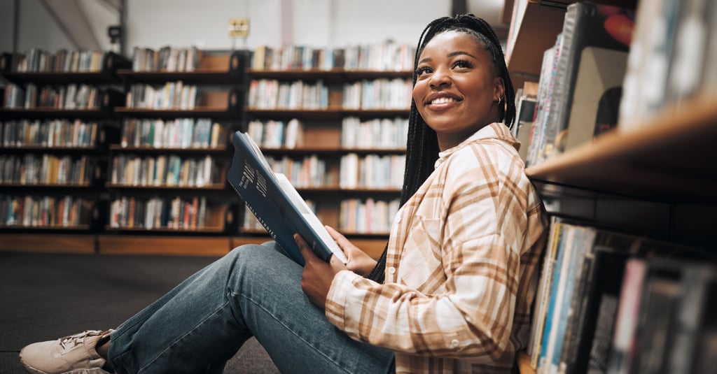 A young woman sits on the floor of a library, smiling and holding an open book. Surrounded by shelves of books, she appears relaxed and happy, perhaps enjoying her studies thanks to financial aid support.
