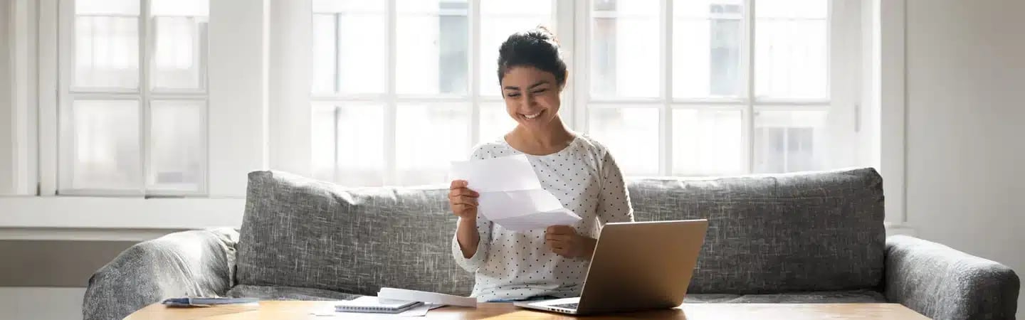 A person sitting on a sofa, smiling while reading a paper, with a laptop, notebook, and pen on the table in front.