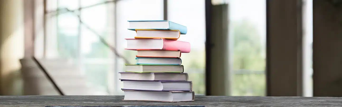 A stack of colorful books sits on a wooden table with a blurred background of large windows and a staircase.