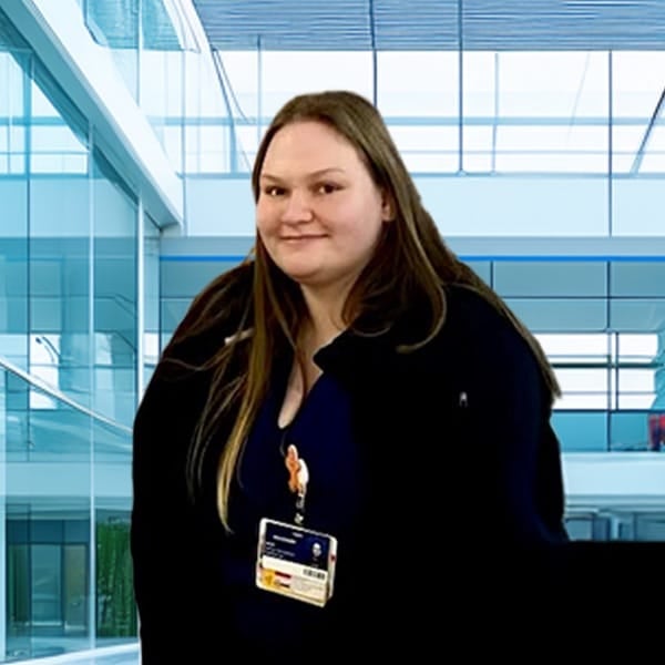 A woman with long brown hair wearing a dark jacket and an ID badge stands indoors in a modern, bright office building with large windows.