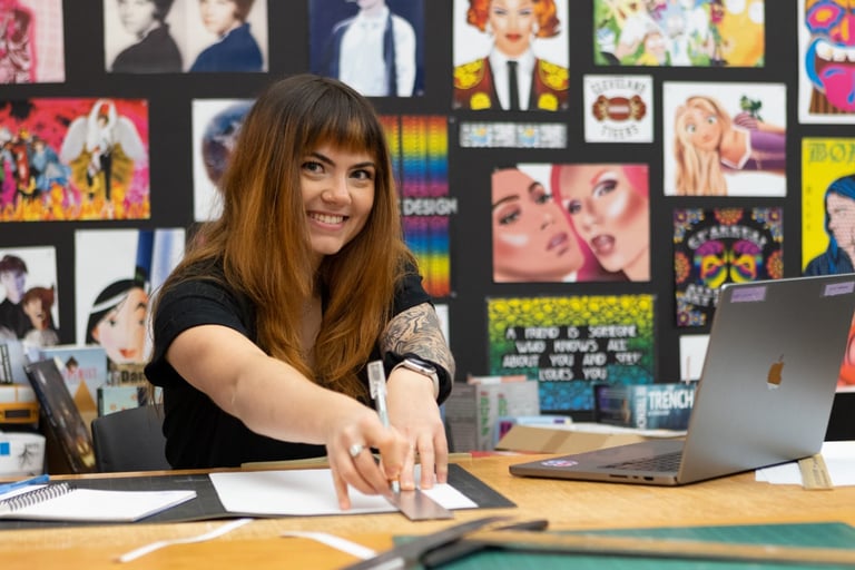 A person smiles while using a ruler at a desk, surrounded by colorful posters and a laptop.