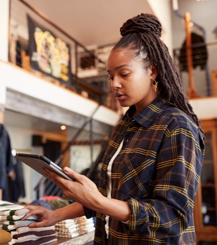 A retail salesperson with long braided hair browses a tablet while standing by neatly folded clothes in a modern clothing store. She appears focused, possibly managing inventory or checking information.