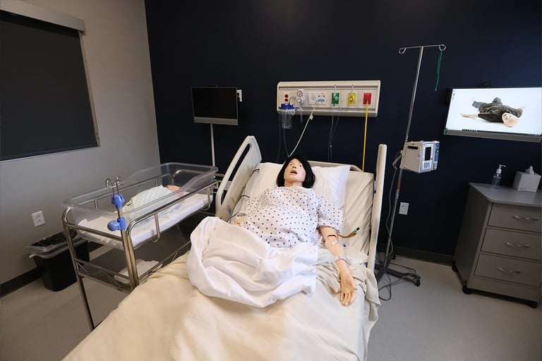 A medical simulation room in Liverpool, NY, features a life-like mannequin in a hospital bed, a baby crib beside it, medical equipment on the wall, and a monitor and IV stand nearby.