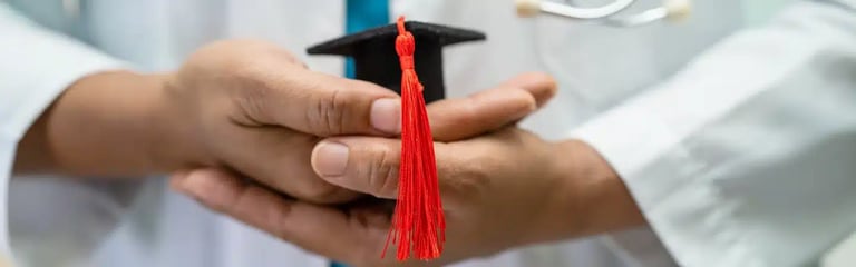 Person in a white coat holding a small graduation cap with a red tassel in their hands.