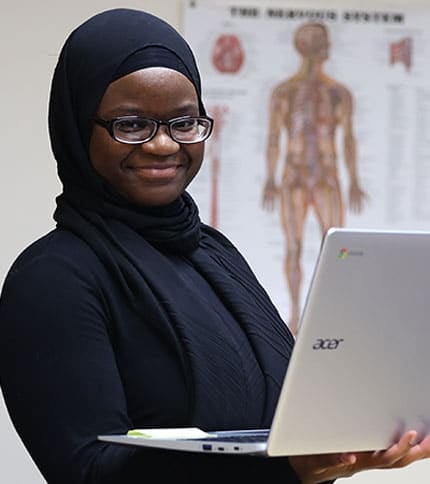 A woman wearing glasses and a headscarf is smiling while holding an Acer laptop. She stands in front of a medical diagram, possibly of the nervous system, on the wall behind her, illustrating her enthusiasm as she embarks on a medical administrative assistant program.