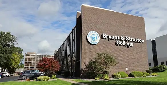 Brick building with a Bryant & Stratton College sign, surrounded by trees and parked cars on a sunny day.