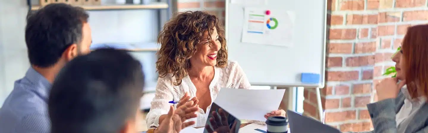 A woman smiling and holding a paper sits at a table with colleagues during a meeting in a bright office.