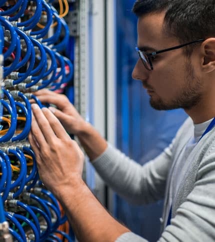 A person wearing glasses and a gray sweater works on a server rack, adjusting blue cables. The background shows more cables and server equipment, indicating a technical environment like a data center.