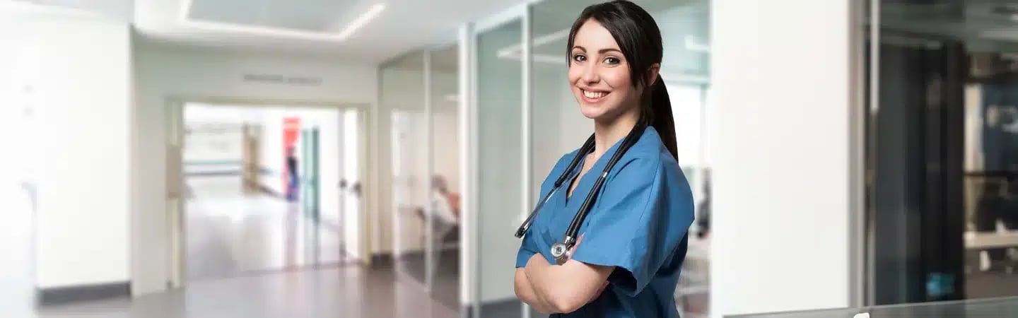 Smiling healthcare worker in blue scrubs with a stethoscope, standing in a hospital hallway with arms crossed.