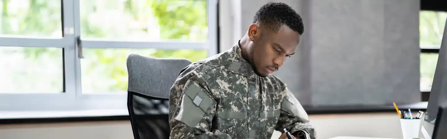 A person in military uniform sits at a desk, focusing on writing or drawing with a pen, in a well-lit office.