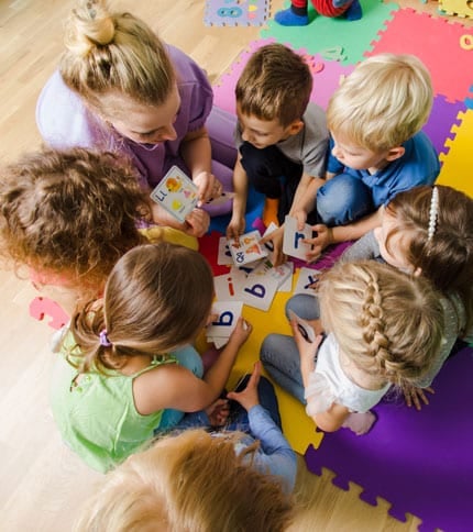 A group of children and a teacher sit on colorful foam tiles in a circle, engaged in a learning activity with alphabet flashcards. The children are focused and interacting with the cards, which are spread out in the center.