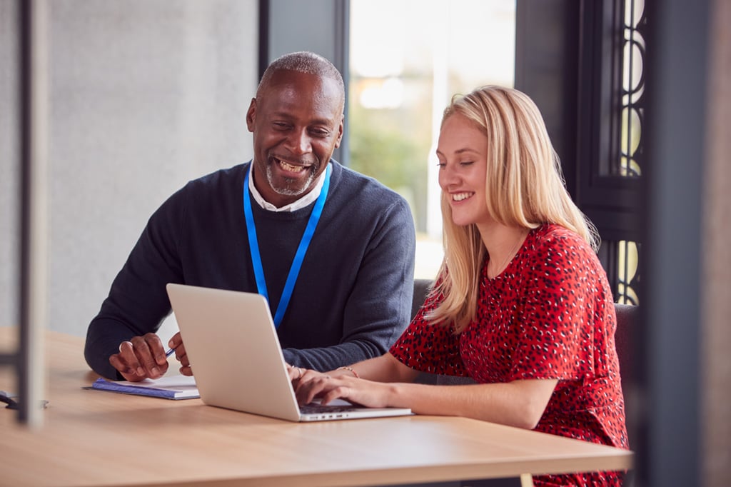 Two colleagues, a man and a woman, sit at a desk smiling as they look at a laptop screen together in a bright office setting, exploring top business degrees to advance their careers.