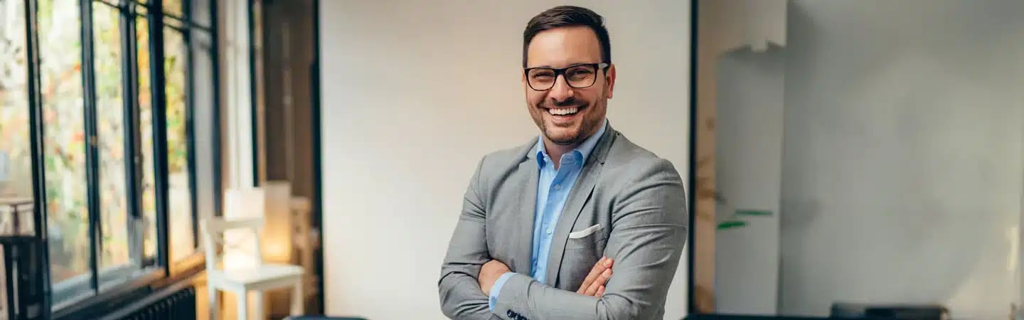A smiling man wearing glasses and a gray blazer stands with his arms crossed in a bright office space.