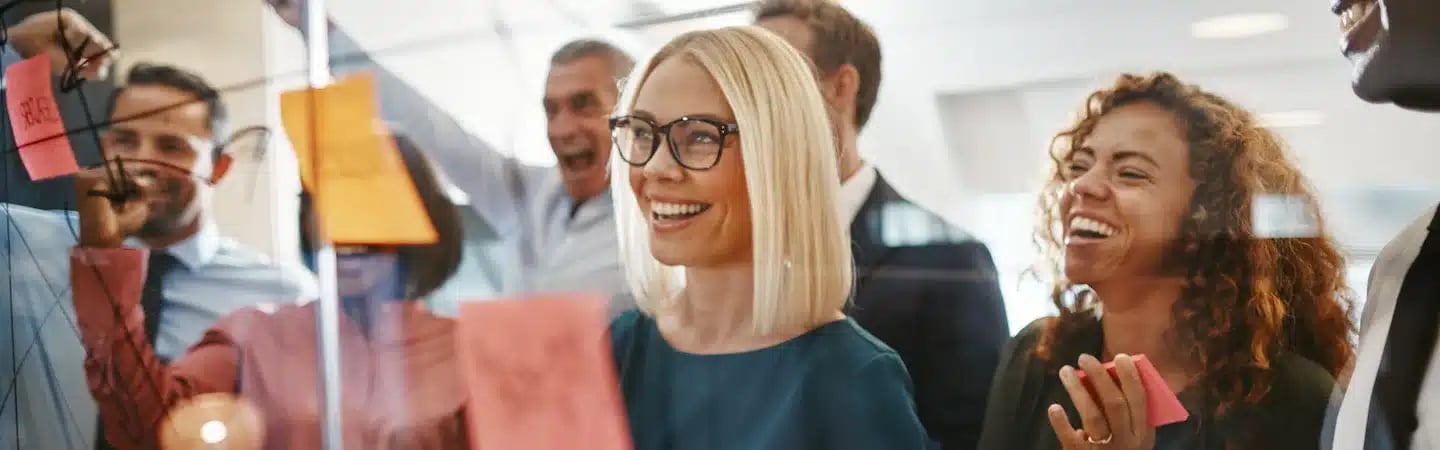 A group of smiling people brainstorm with sticky notes on a glass wall in a bright office space.