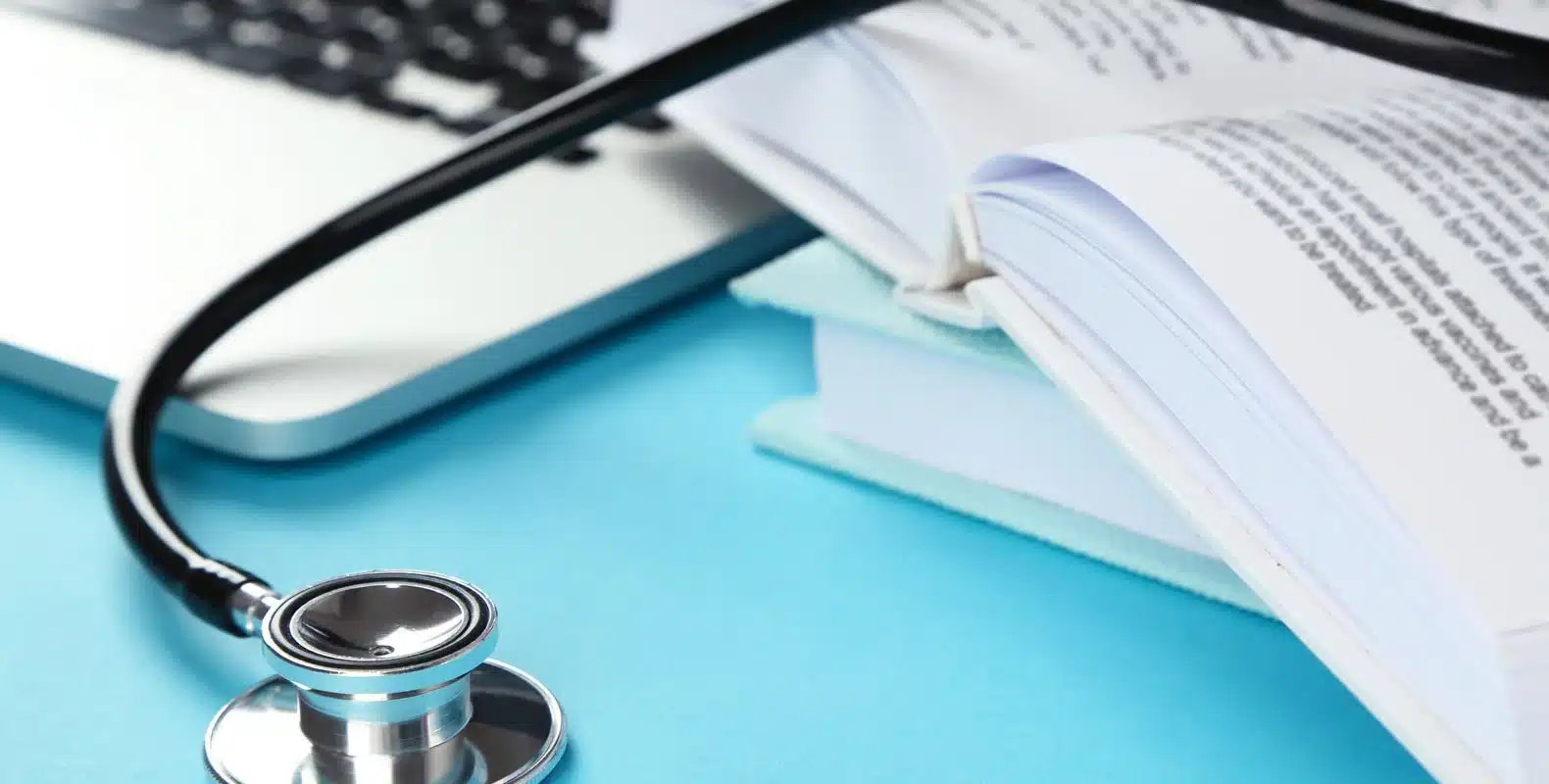 A stethoscope, open book, and a laptop on a turquoise surface.