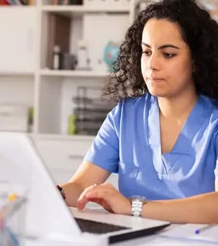 A person with curly hair wearing blue scrubs is sitting at a desk, focused on working on a laptop, possibly studying for an online medical office assistant certification. The background shows bookshelves and office supplies.