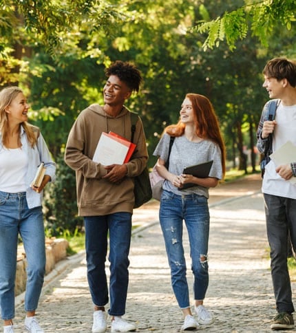Four young adults walk on a tree-lined path, smiling and carrying books. They are casually dressed and appear to be engaged in conversation, enjoying a sunny day outdoors.