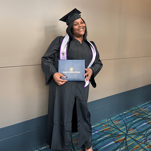 Person in graduation cap and gown, smiling and holding a diploma with "Bryant & Stratton College" text visible.
