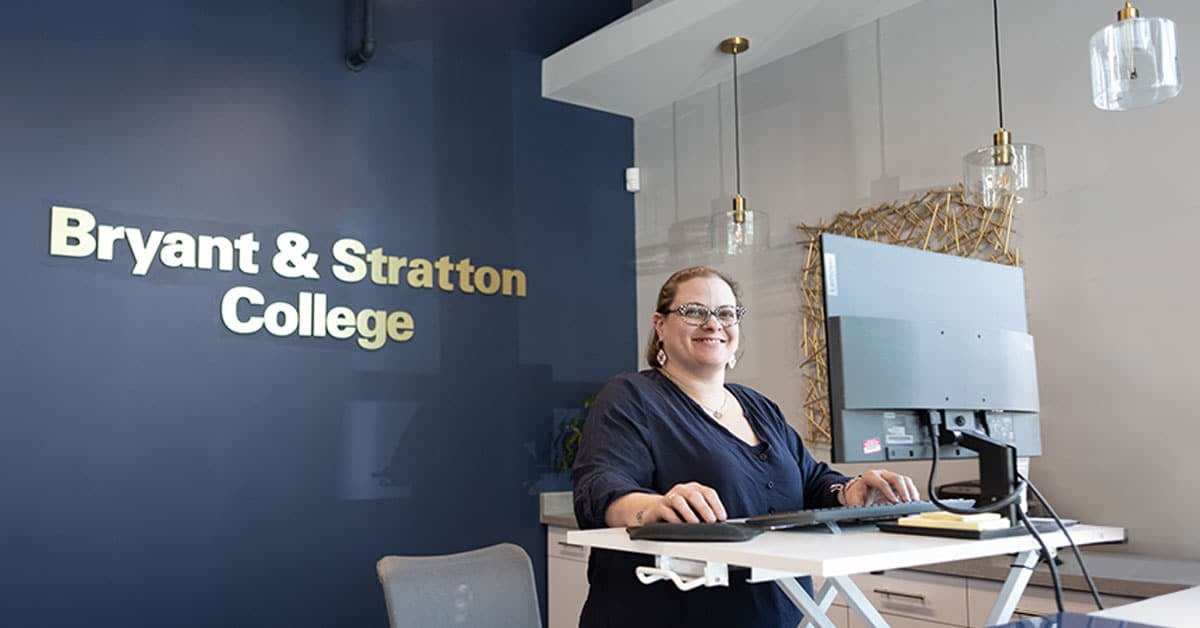 A person stands smiling at a standing desk with a computer. Behind them is a sign that reads "Bryant & Stratton College."
