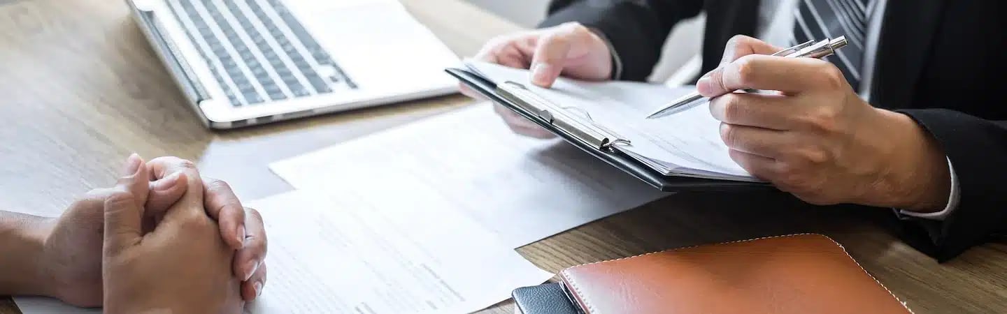 Two people in a business meeting; one holding a clipboard and pen, the other with clasped hands on the table.