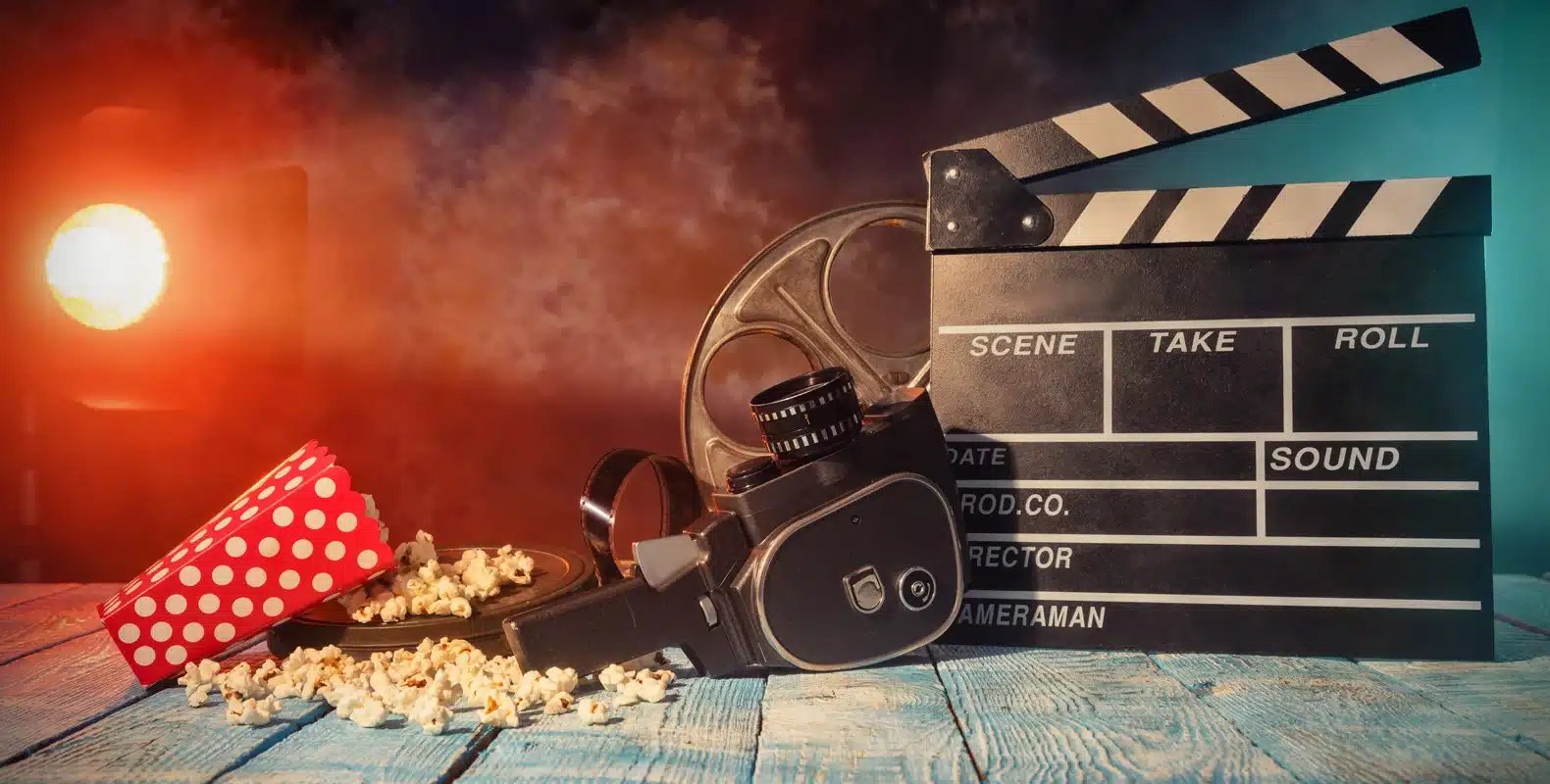 Vintage film camera, popcorn, film reel, and clapperboard on a wooden table with cinematic lighting in the background.