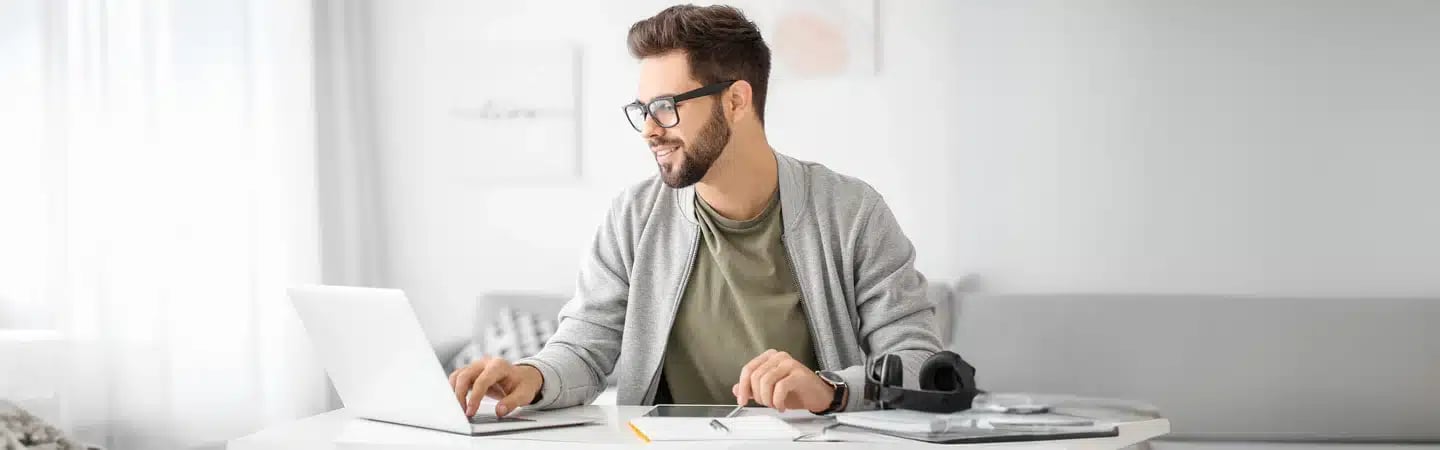 A man with glasses, wearing a gray cardigan, works on a laptop at a white desk with papers and a tablet.