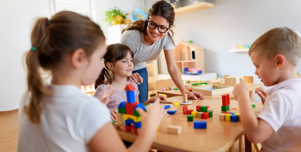 A woman with a CDA Credential smiles as she supervises three young children playing with colorful wooden blocks at a table in a bright, cheerful early childhood education classroom.