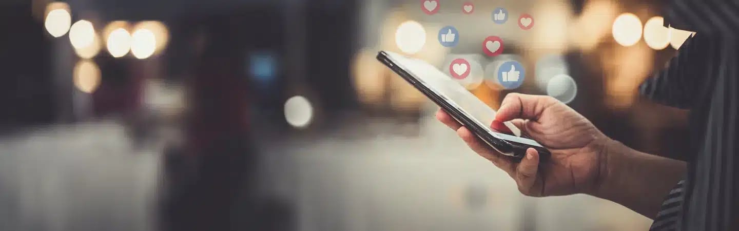 Close-up of a person holding a smartphone with floating Facebook-like thumbs-up and heart icons on a blurred background.