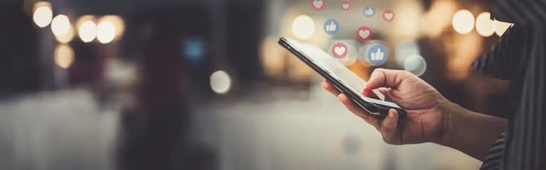 Close-up of a person holding a smartphone with floating Facebook-like thumbs-up and heart icons on a blurred background.