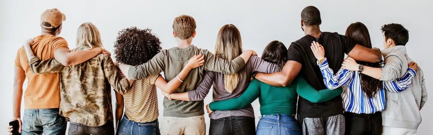 A diverse group of people stand in a row with their backs turned, arms around each other in a show of unity and friendship.