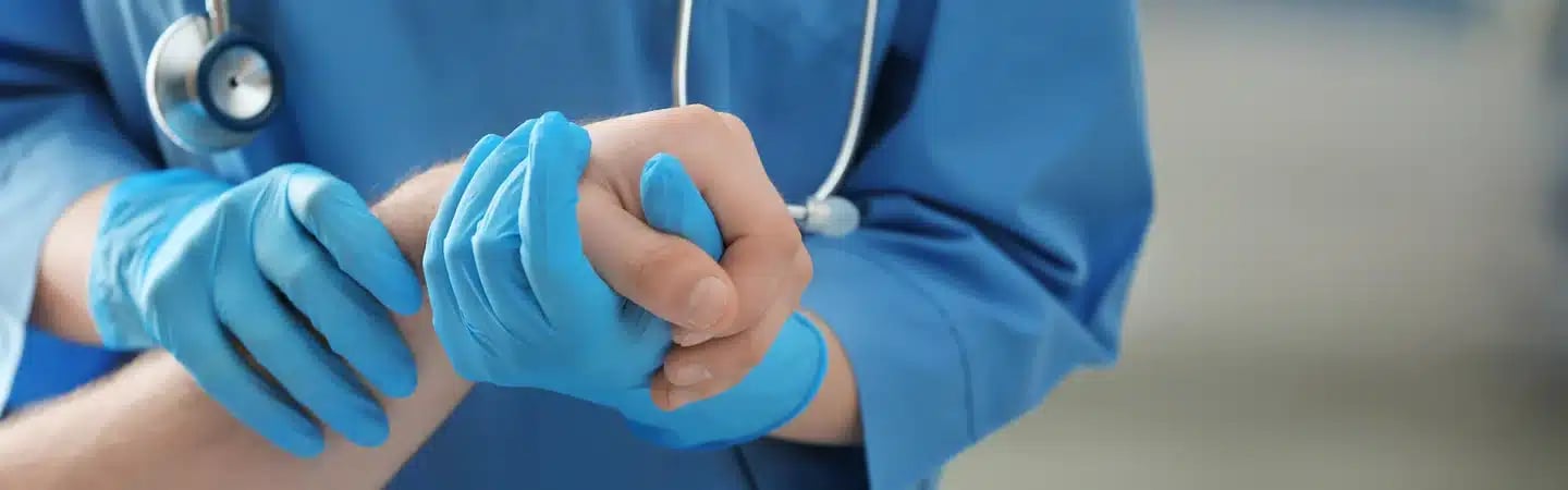 Close-up of a healthcare professional in blue scrubs and gloves checking the pulse of a patient's wrist.