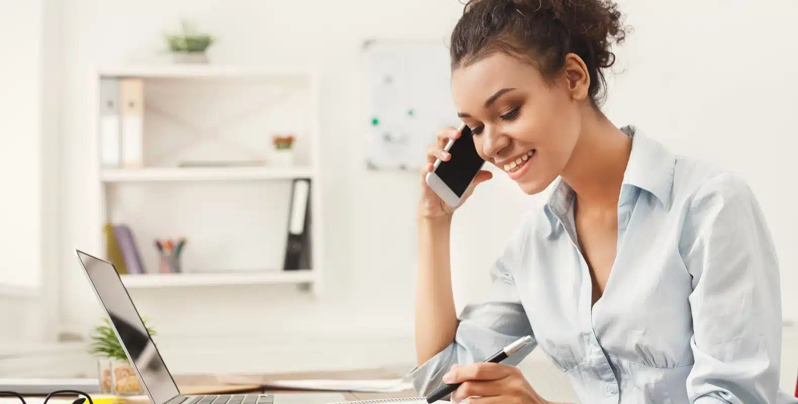 Woman smiling at desk, talking on phone, writing, with laptop open in bright office.